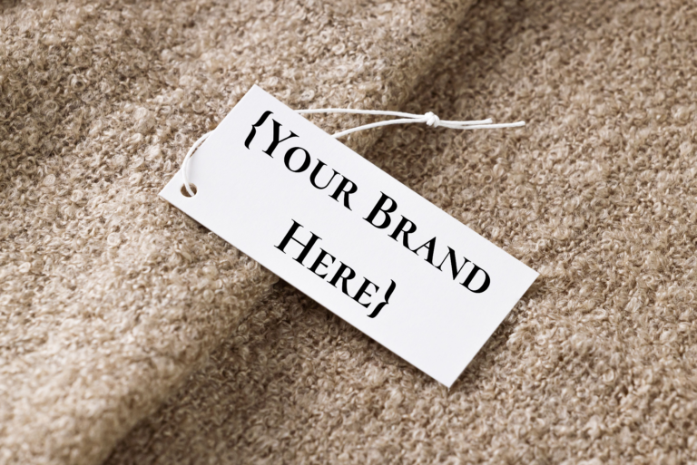 Picture is a branding tag with a string sitting on a piece of tan fabric. Overlayed the tag are the words "{Your Brand Here}" reflecting that branded items are important