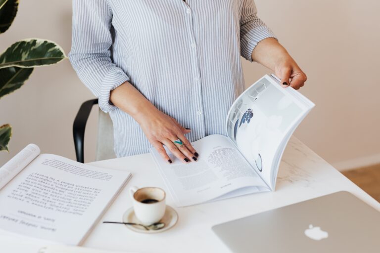 a woman standing at a table with a coffee mug, laptop, and guidebook. She is looking through the guidebook