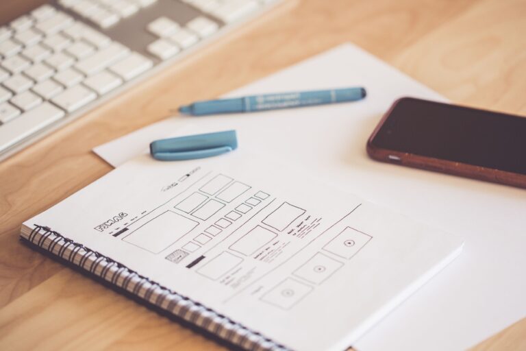 image shows a website wireframe sketched onto a piece of paper, with a pen and phone nearby, all sitting on a light oak desk
